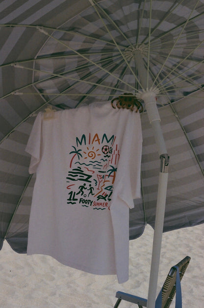White 1Language Endless Summer Tee hanging beneath a beach umbrella with its back graphic visible