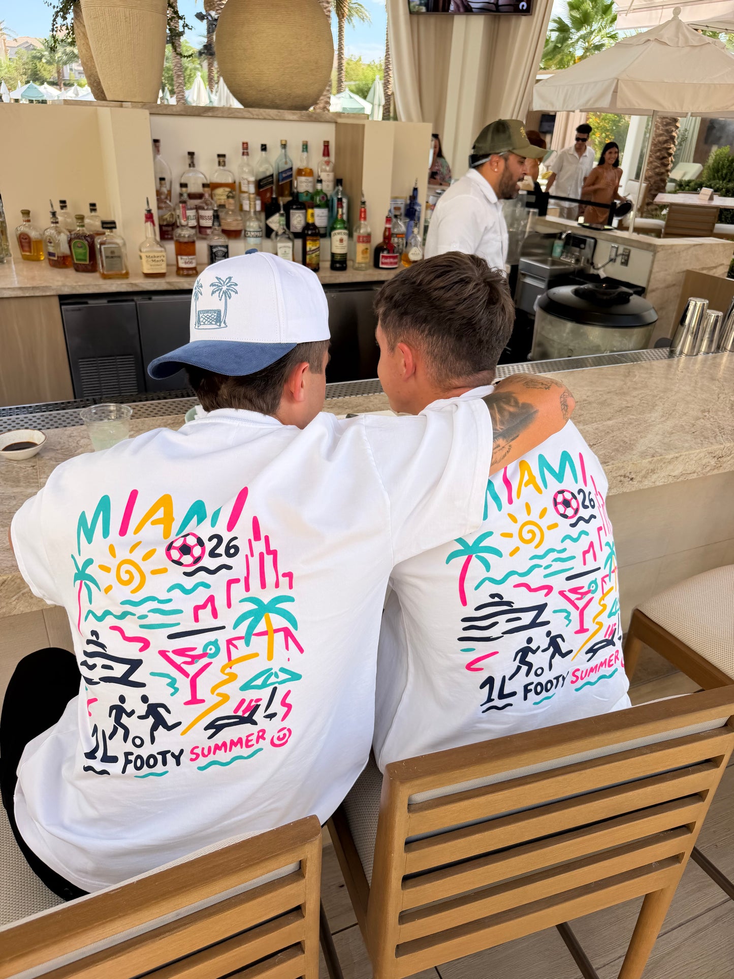 Two friends wearing white 1Language Endless Summer graphic T-shirts at a poolside bar in Miami