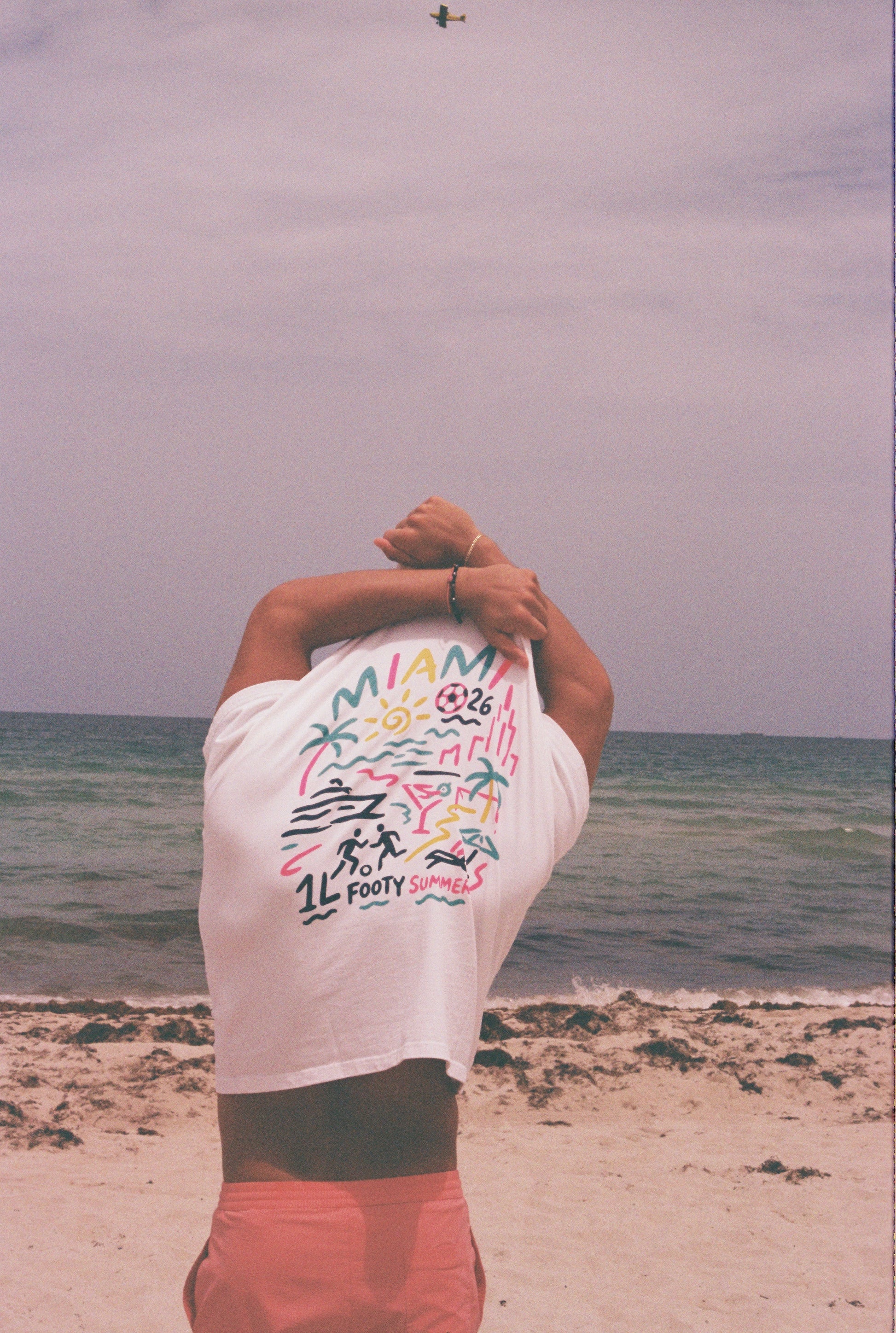 Model showing the colorful Miami-inspired back graphic of the white Endless Summer Tee at the beach